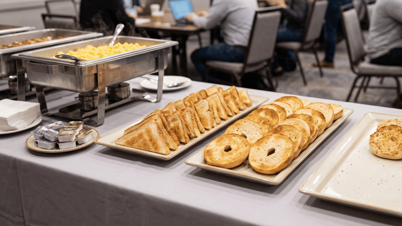Buffet-style breakfast setup with trays of toasted bread slices, bagels, and English muffins arranged on a table beside a chafing dish in a dining area, ready for self-serve guests.